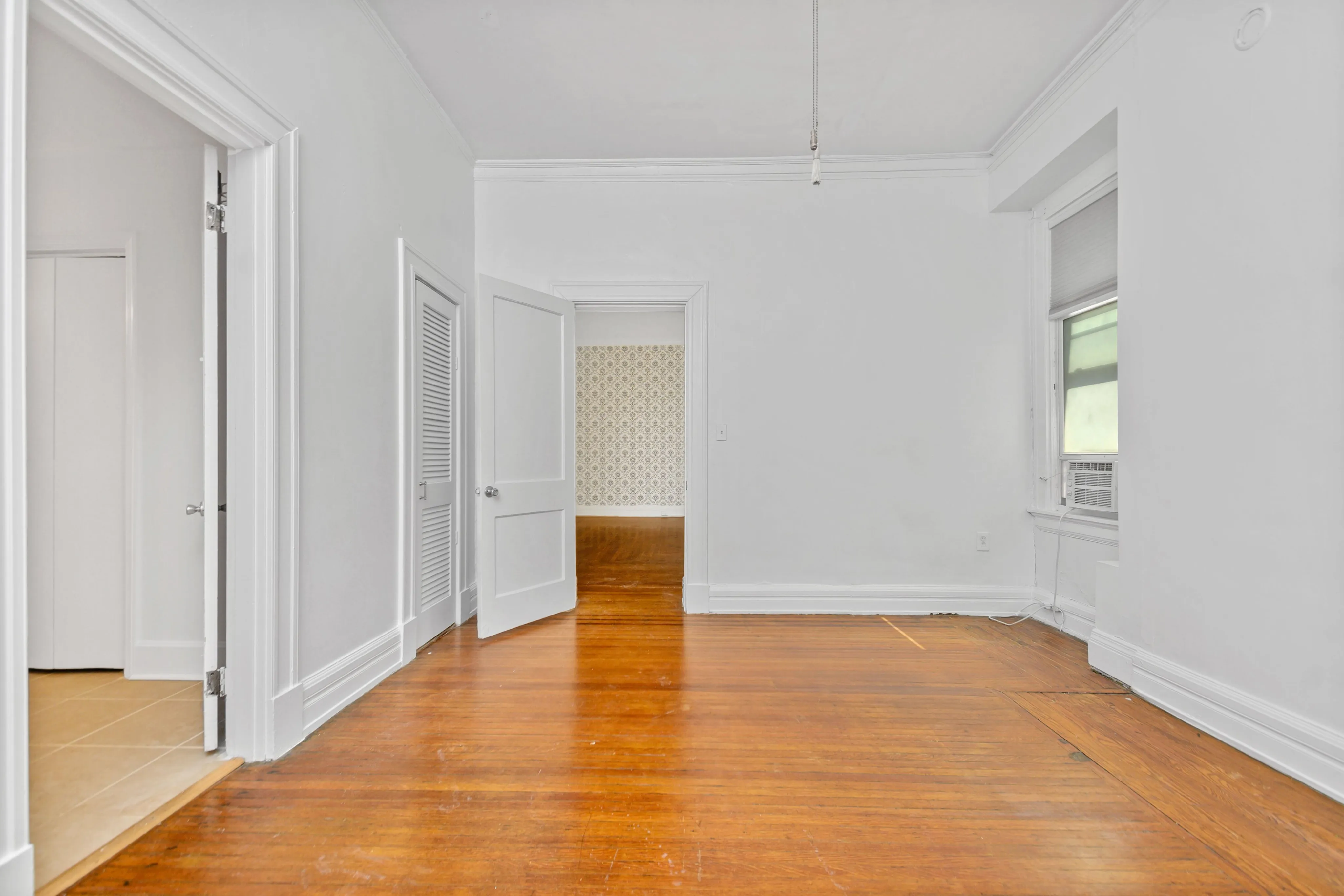 Bright and spacious room with elegant hardwood floors, white walls, and an open doorway revealing patterned wallpaper in the next room.