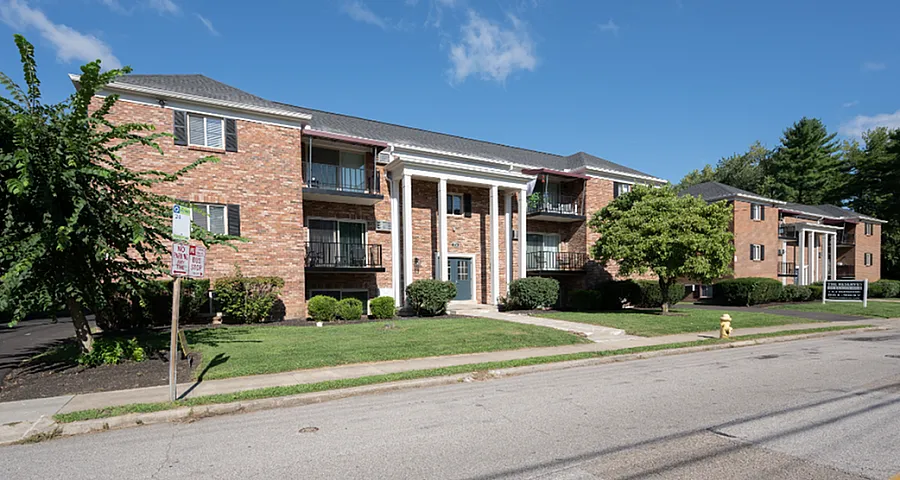 Charming brick apartment building with elegant white columns, surrounded by lush greenery and a clear blue sky.