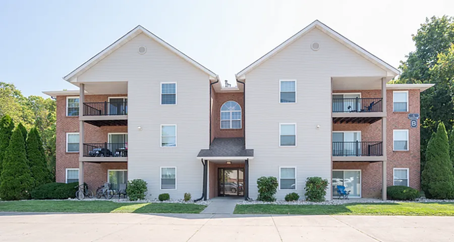 Charming three-story brick apartment building with spacious balconies, surrounded by lush greenery and a well-maintained pathway.