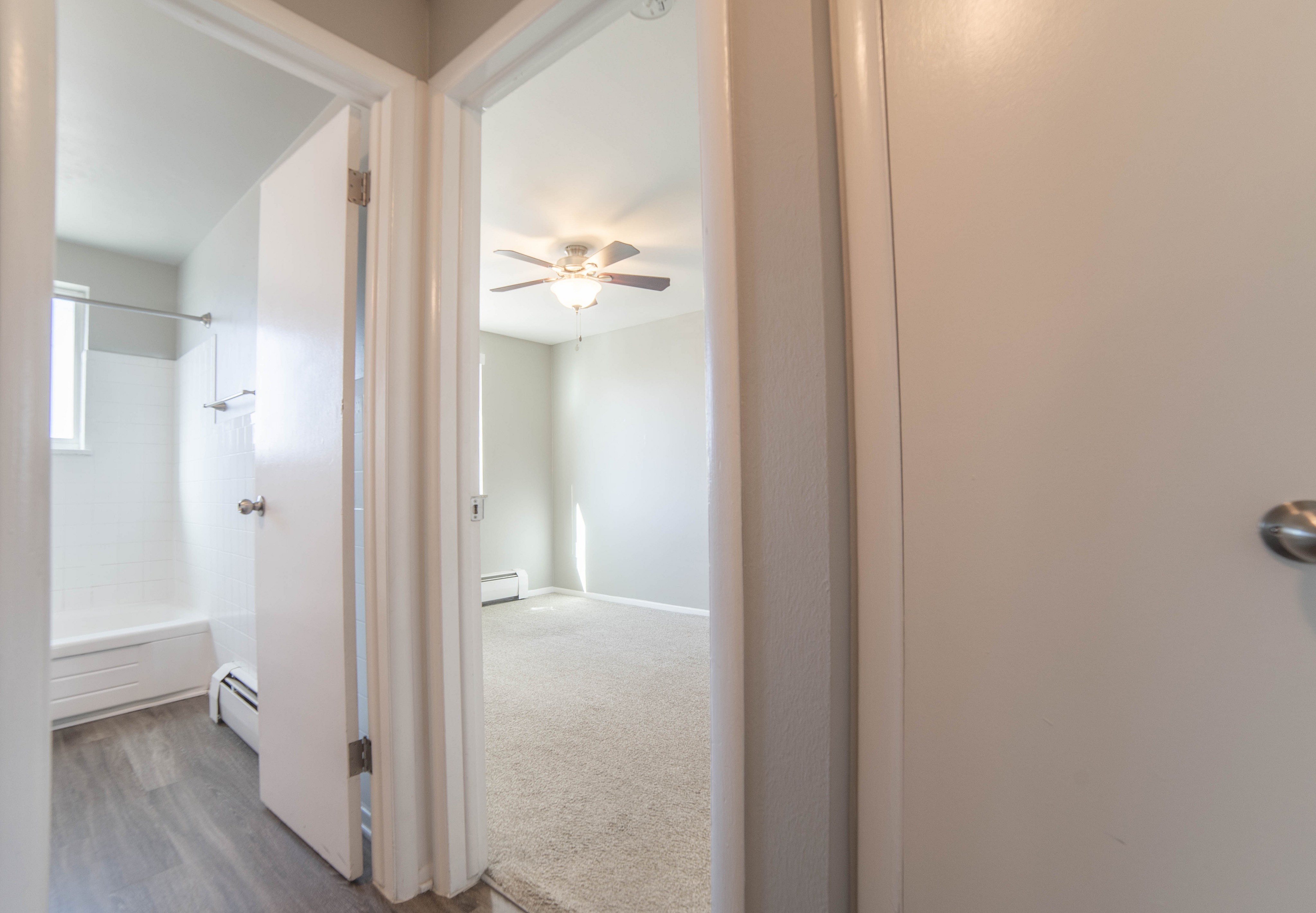 Inviting hallway view showcasing a bright and airy bedroom with soft carpet and a ceiling fan, adjacent to a modern bathroom with sleek floors.