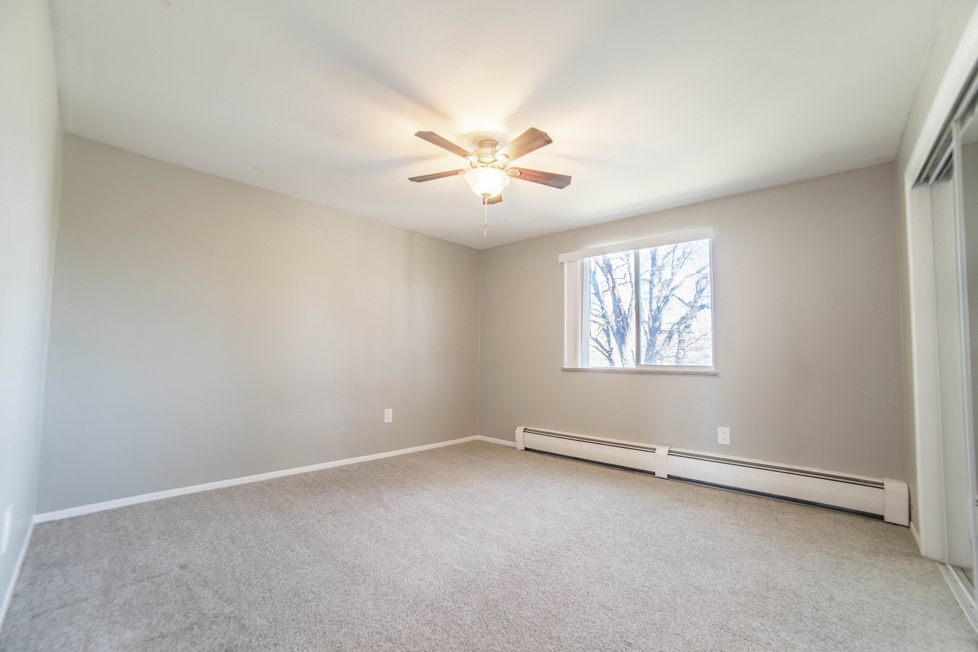 Bright and spacious bedroom with plush carpeting and a ceiling fan, featuring a large window with a serene view.