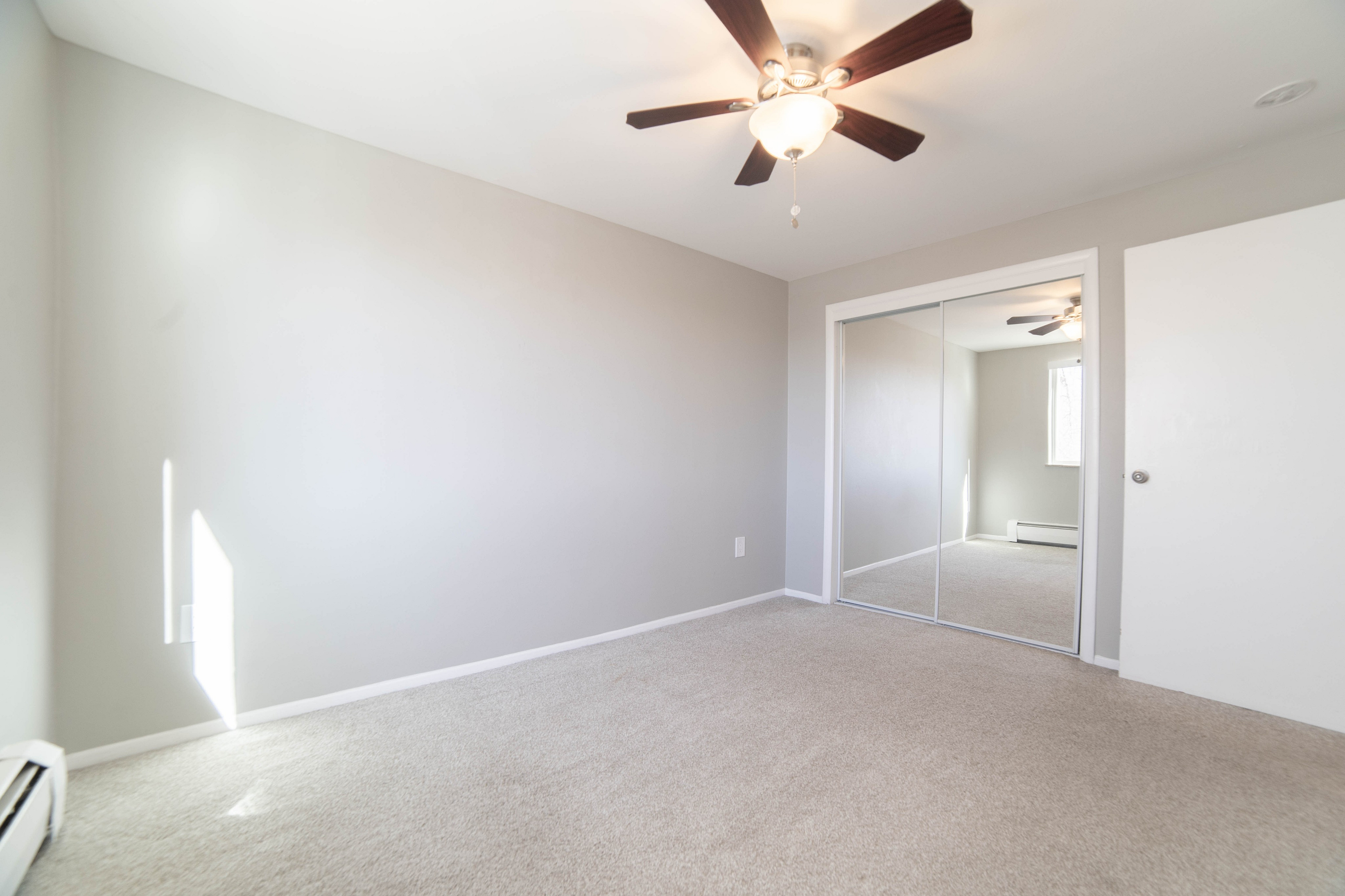 Bright and airy bedroom with plush carpeting, mirrored closet doors, and a modern ceiling fan.