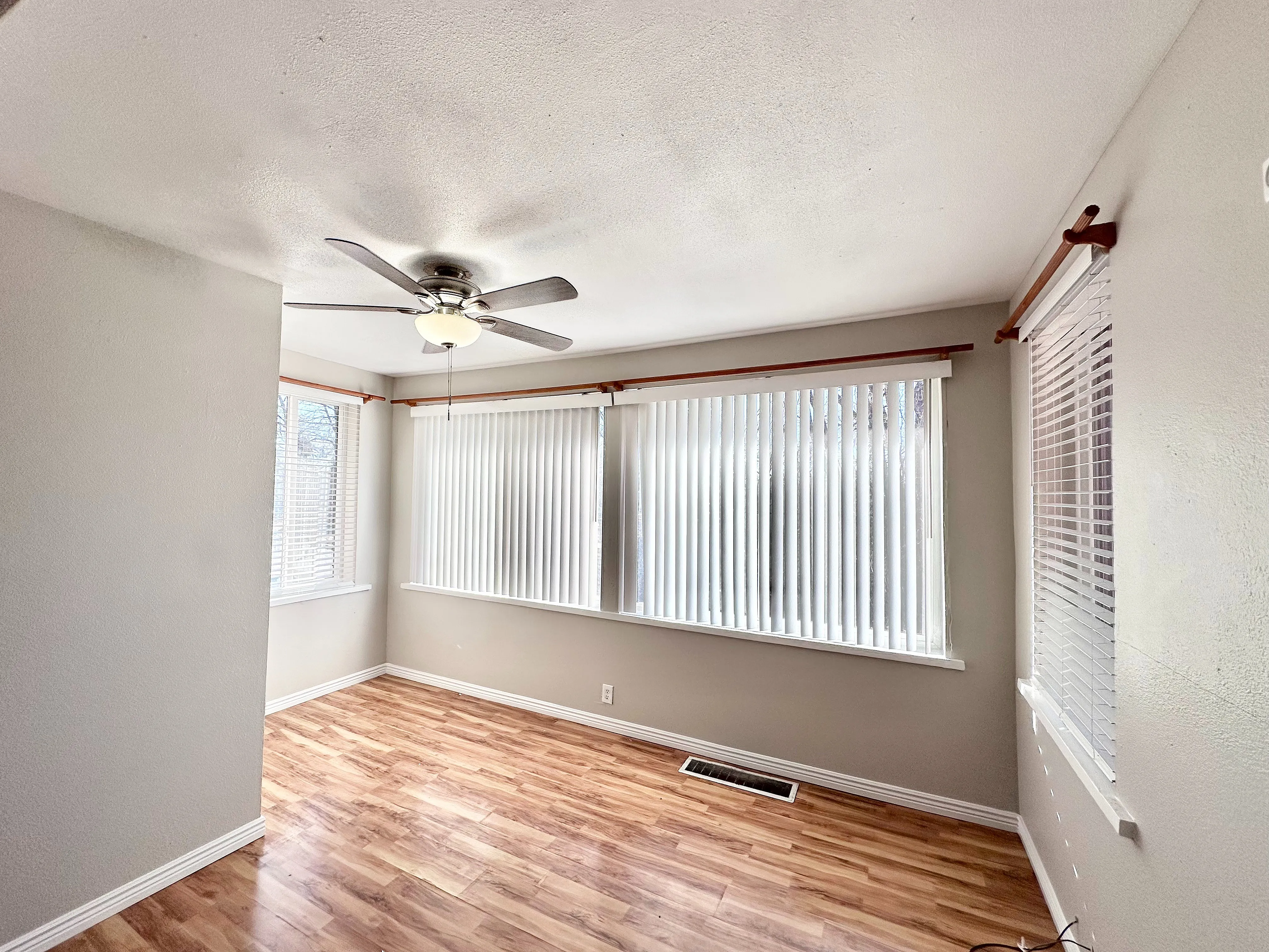 Bright and airy room boasting elegant hardwood floors, large windows with vertical blinds, and a stylish ceiling fan.