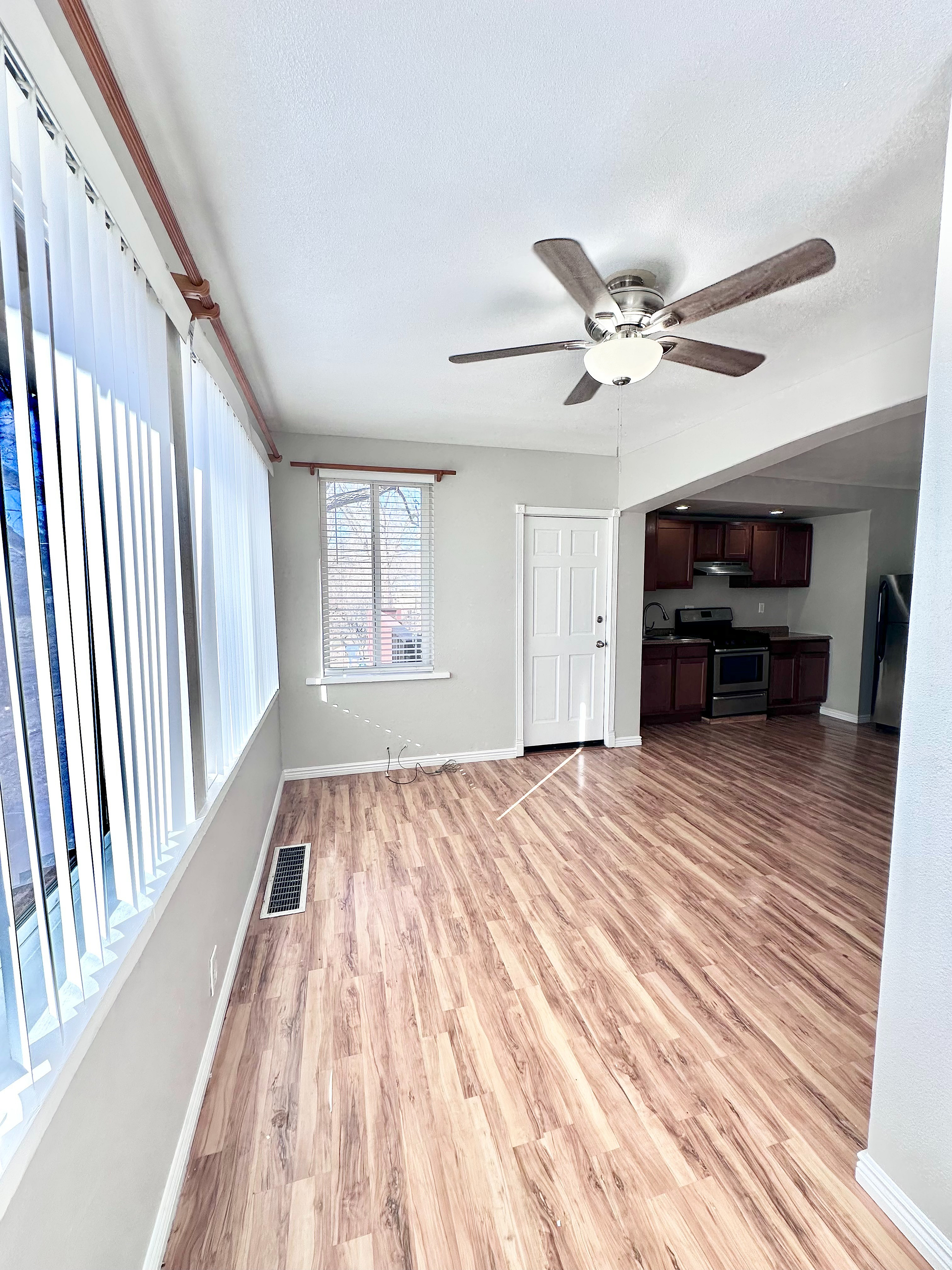 Bright and inviting living area with elegant wood flooring and modern ceiling fan, featuring large windows with vertical blinds in a contemporary open layout.
