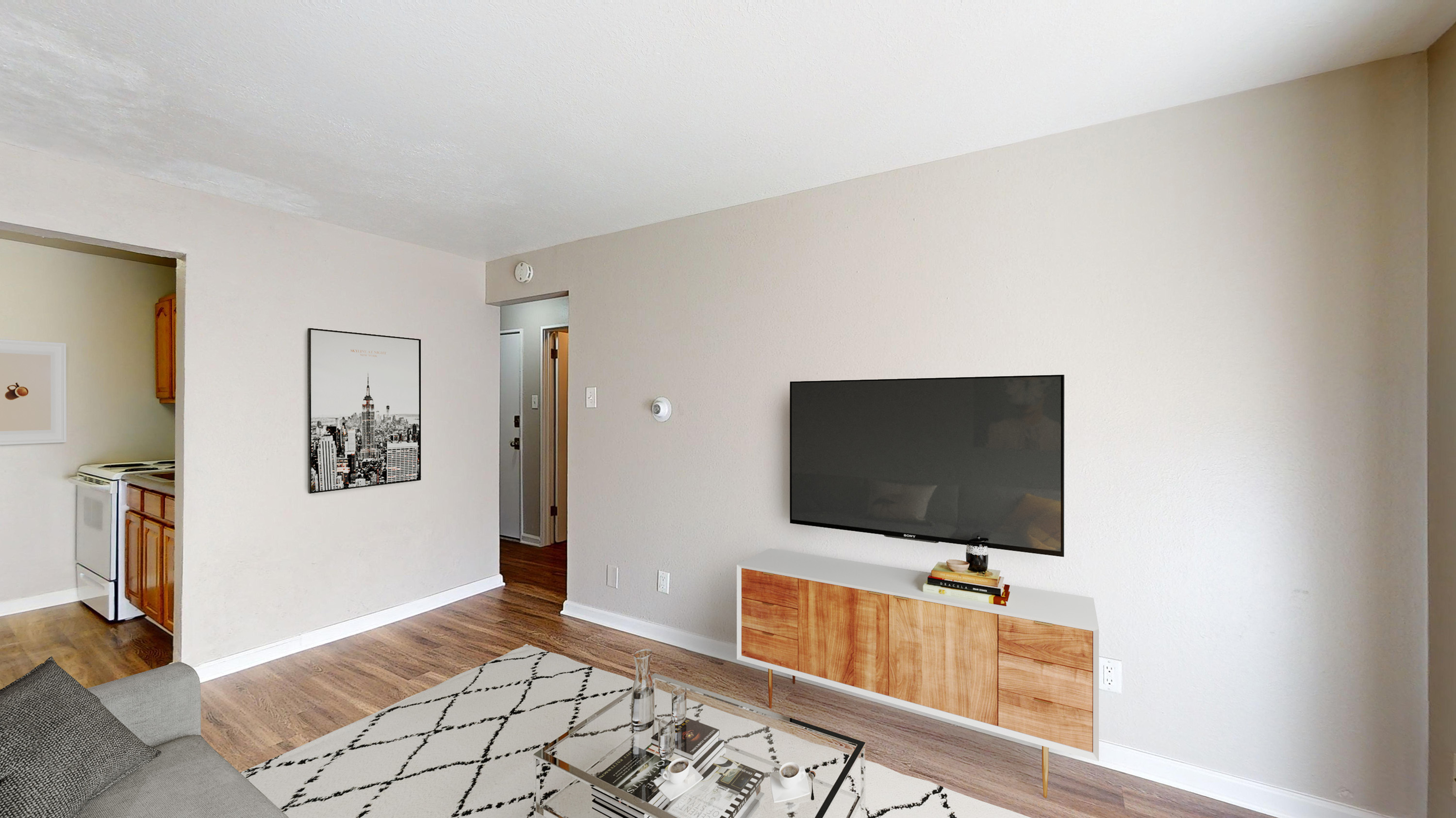 Modern living room with natural light, sleek wooden flooring, and a stylish TV unit. Adjacent kitchen area with classic cabinetry visible.