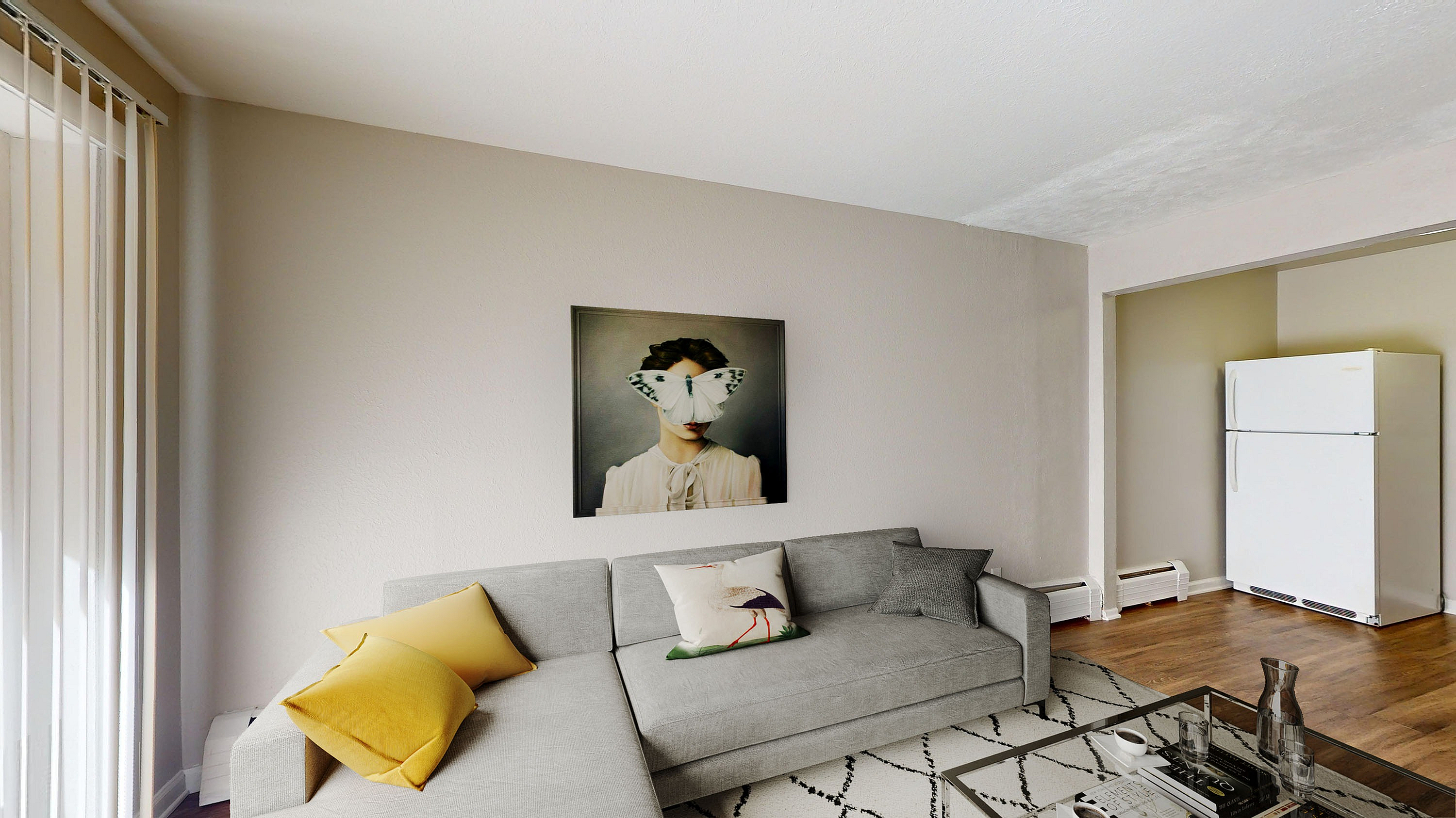 Modern living room with a stylish gray sofa adorned with vibrant yellow and patterned cushions, contemporary art on the wall, and an inviting atmosphere near the kitchen area with natural light streaming through vertical blinds.