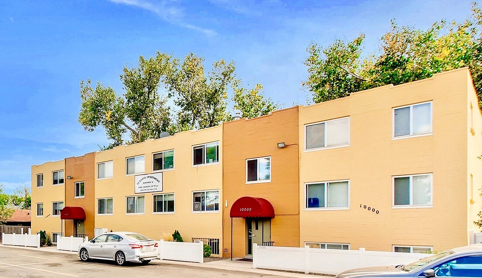 Charming yellow apartment building with red awnings, surrounded by lush trees and spacious parking area.