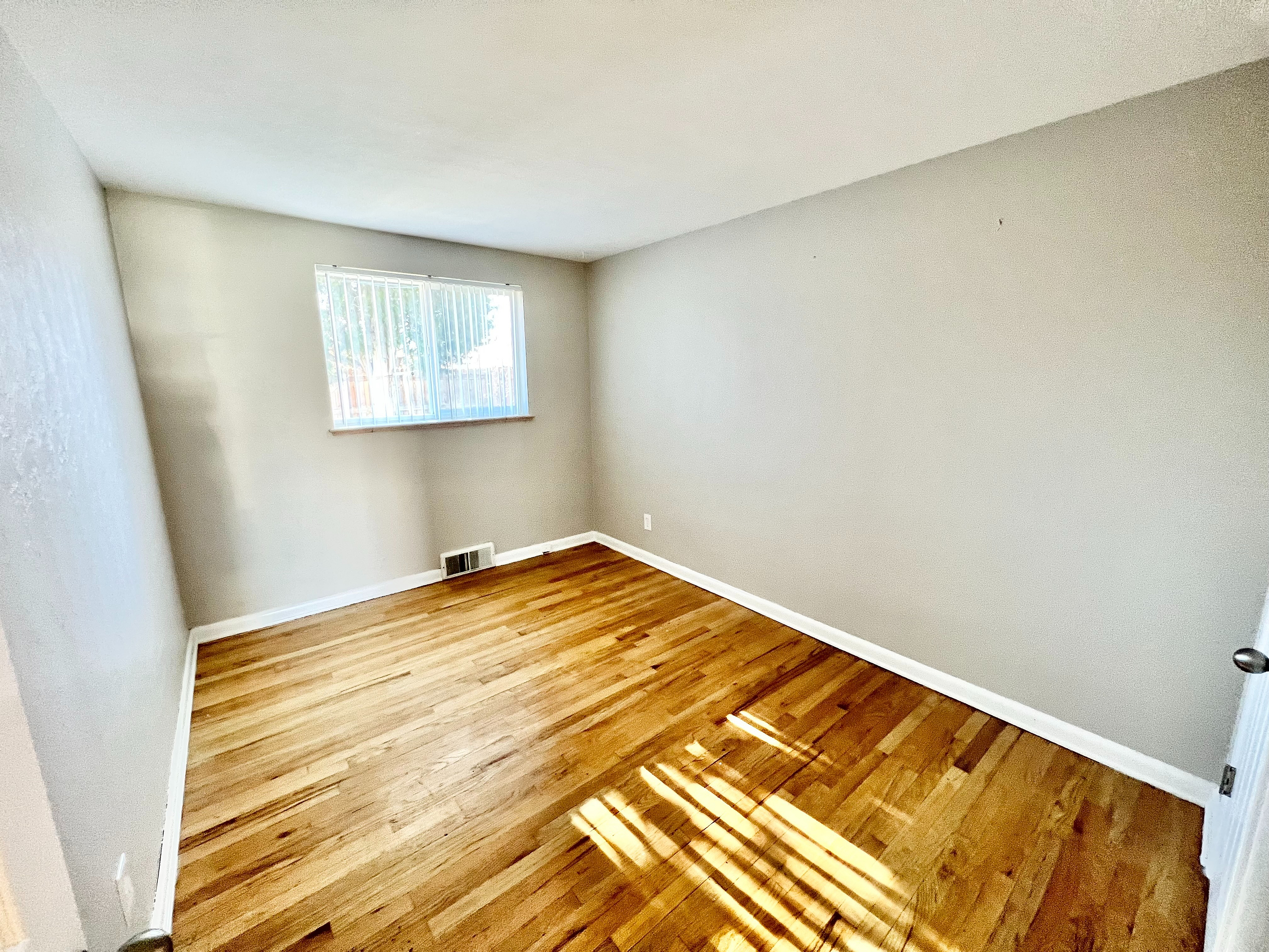 Bright and inviting room with freshly polished hardwood floors and ample natural light from a large window, perfect for a cozy bedroom or home office.