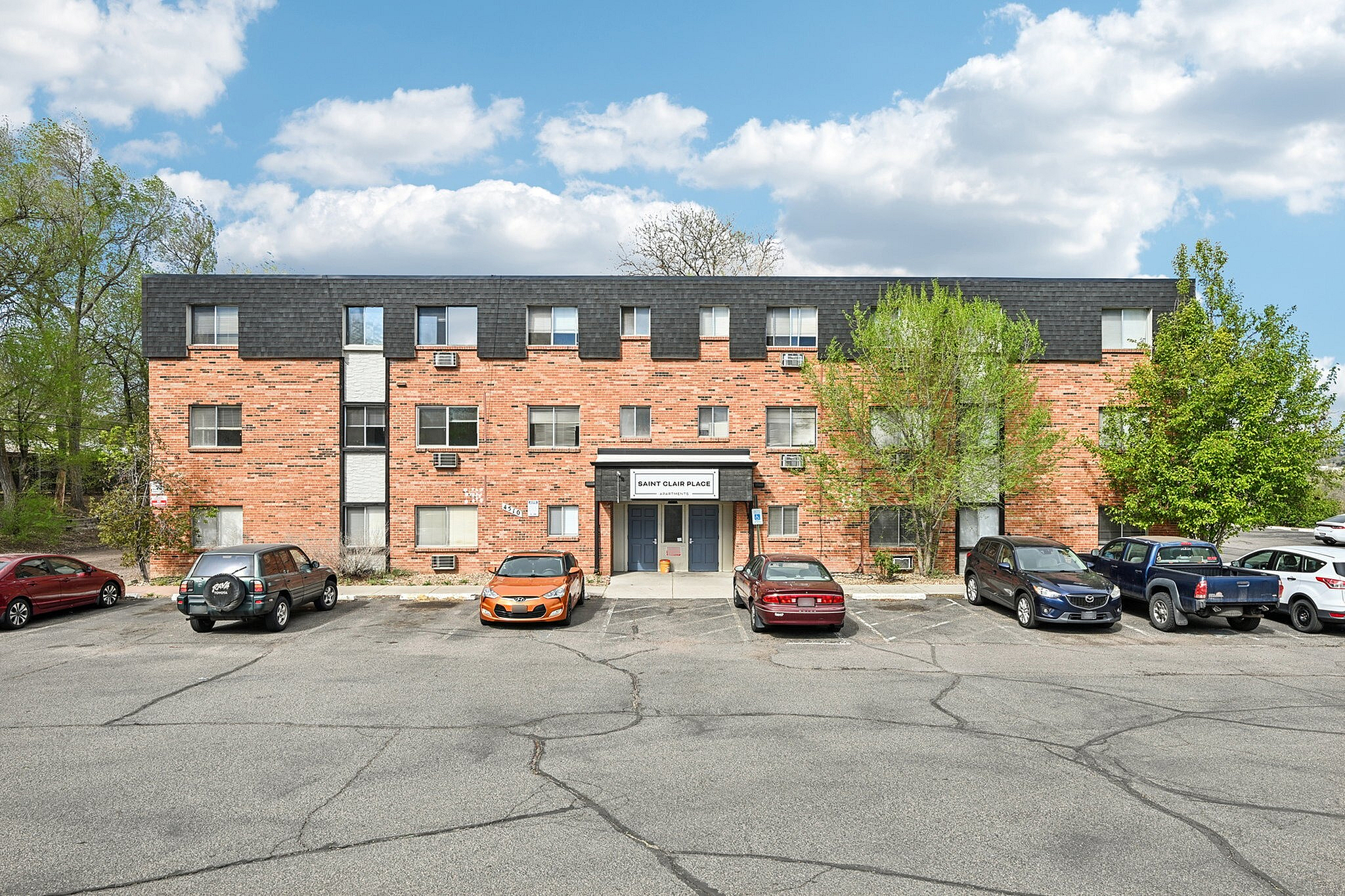 Charming brick apartment building with sleek black roofing, surrounded by lush greenery and ample parking space under a vibrant blue sky.