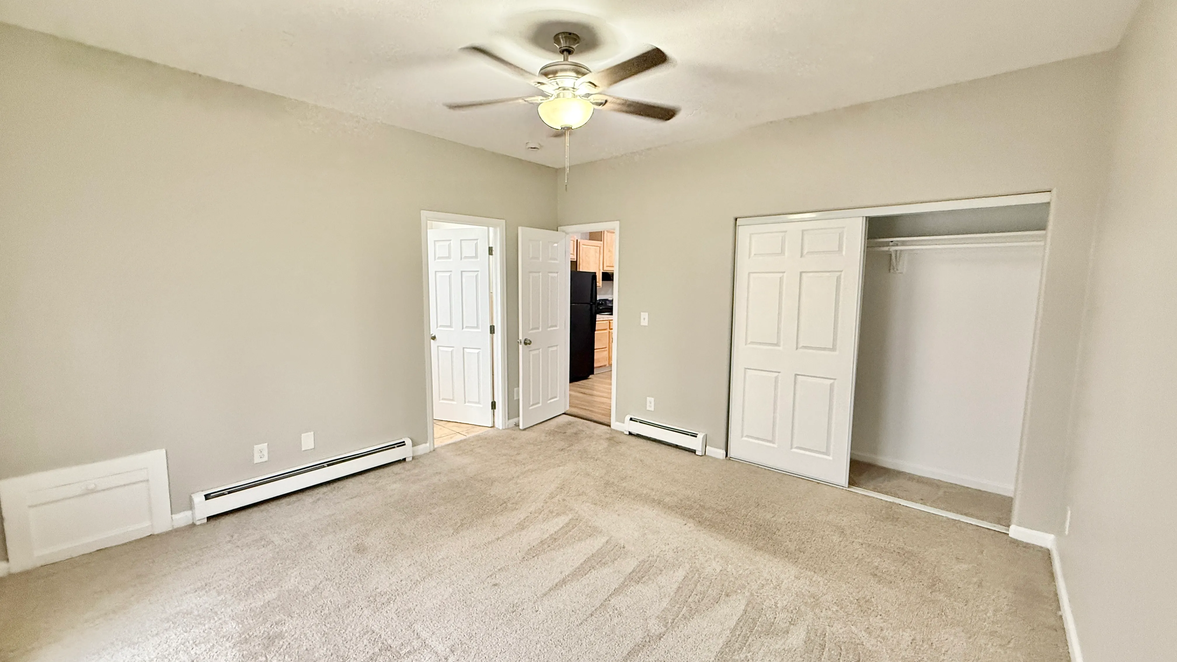 Bright and spacious bedroom featuring plush carpet, a ceiling fan, and ample closet space with double doors, leading into a well-lit hallway.