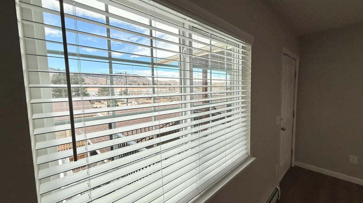 Bright and airy apartment view through large window blinds showcasing scenic mountain landscape.