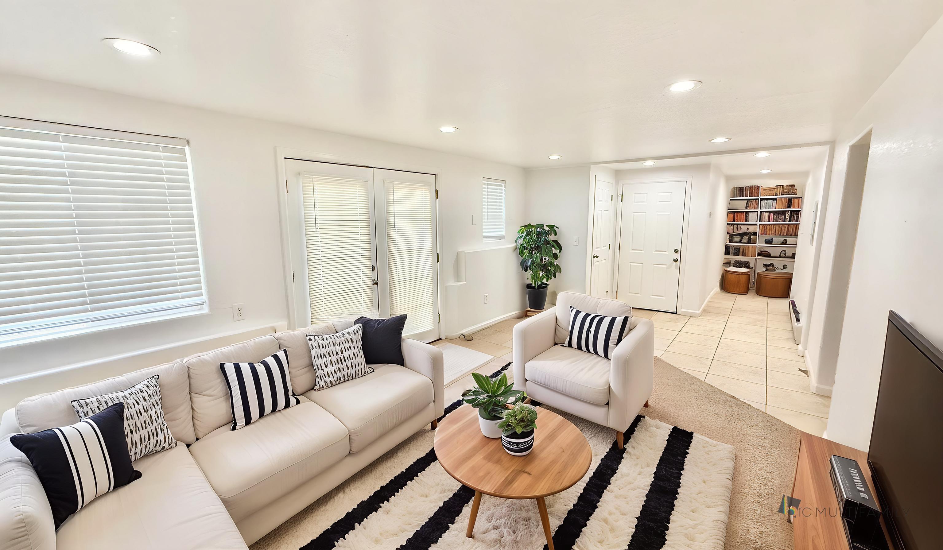 Bright and inviting living room with cozy white sofas, stylish striped cushions, and a chic wooden coffee table adorned with plants, leading to a tiled entryway with built-in shelves.