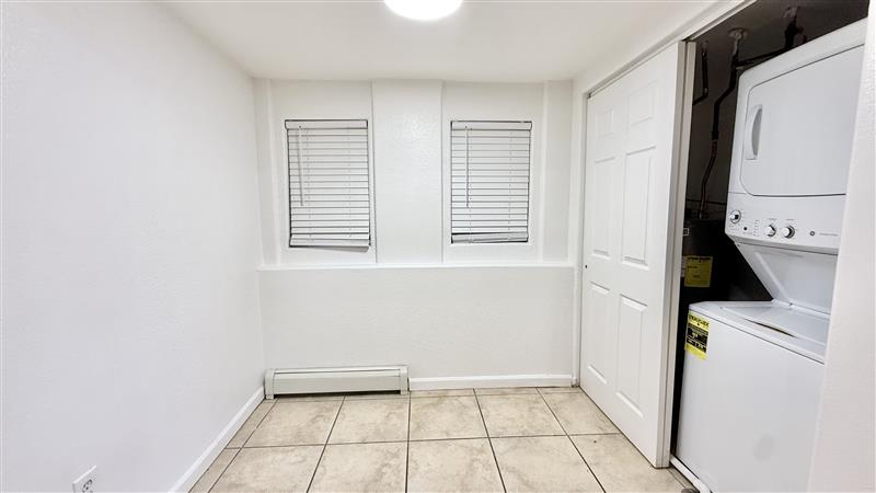 Bright laundry nook with stacked washer and dryer, tiled flooring, and two windows with blinds.