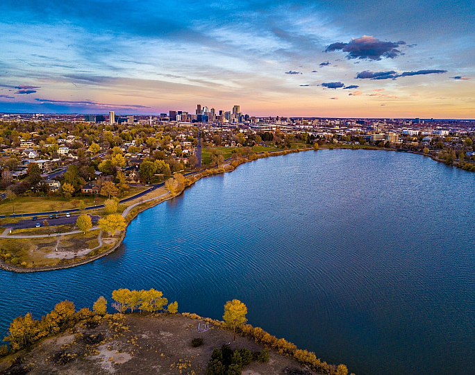 Aerial view of a serene lake with vibrant autumn foliage, adjacent to a bustling cityscape at sunset.