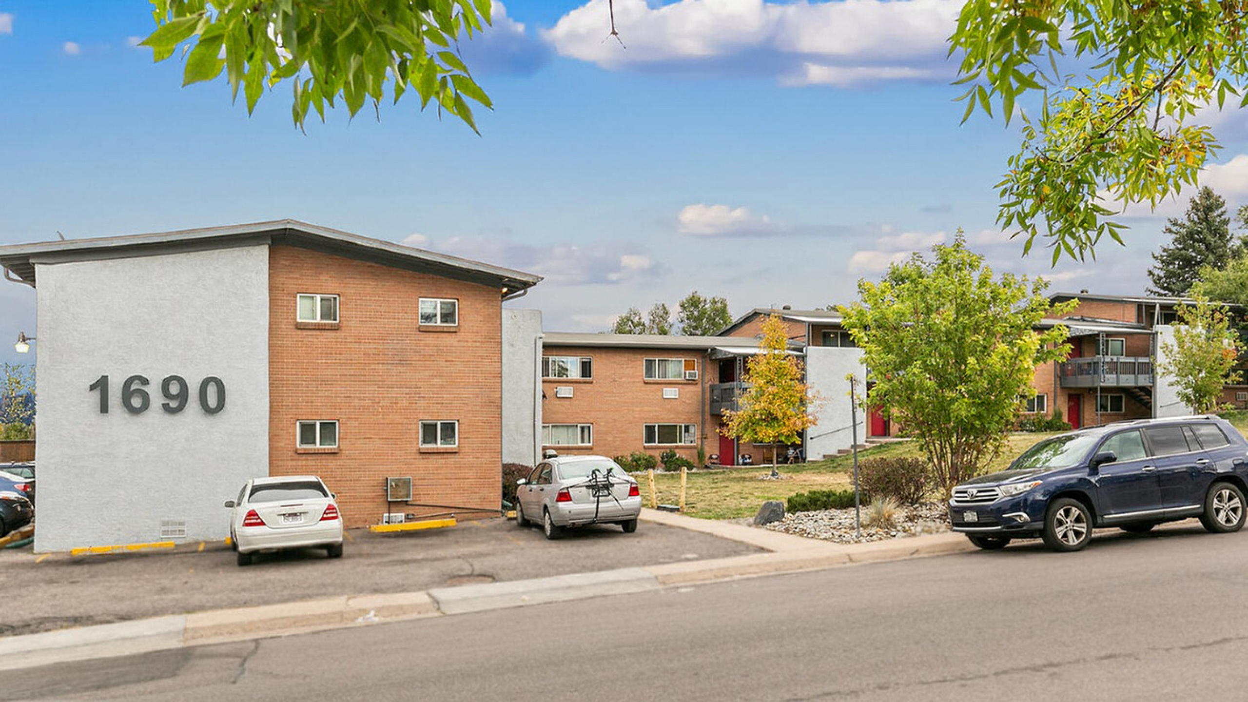 Exterior view of a brick apartment building with lush greenery and parked cars.