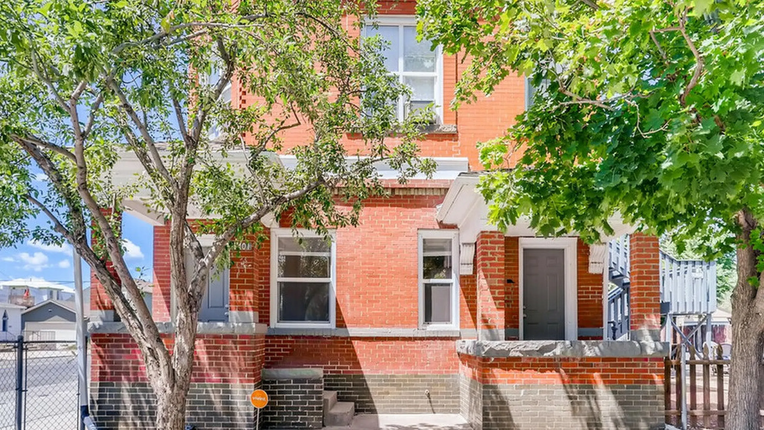 Charming red brick home with lush green trees and a sunny sky background.