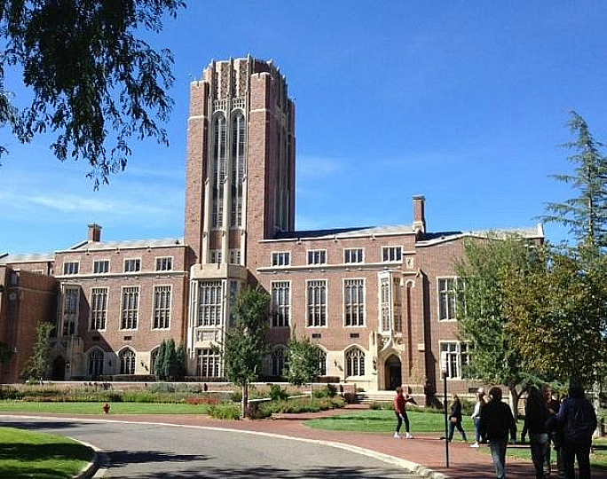 Impressive historic brick building with a tall central tower, surrounded by lush greenery and a clear blue sky, offering an inviting academic atmosphere.