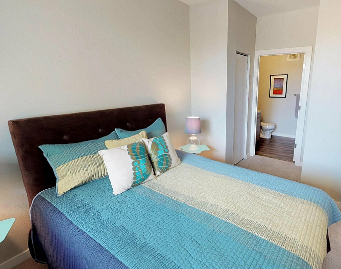 Cozy bedroom with a plush bed adorned in teal and cream linens, colorful pillows, chic side tables, and modern decor leading into a contemporary bathroom.