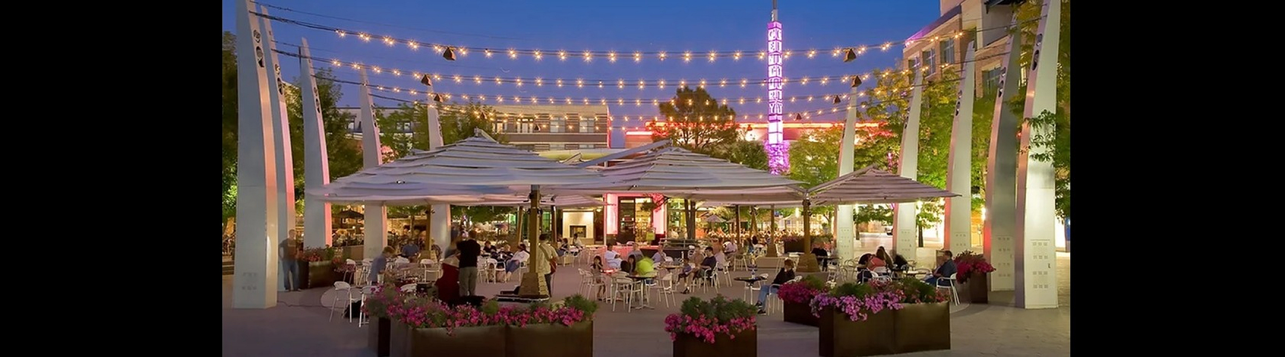 Charming urban outdoor dining area with string lights, vibrant flowers, and a lively atmosphere under the evening sky.