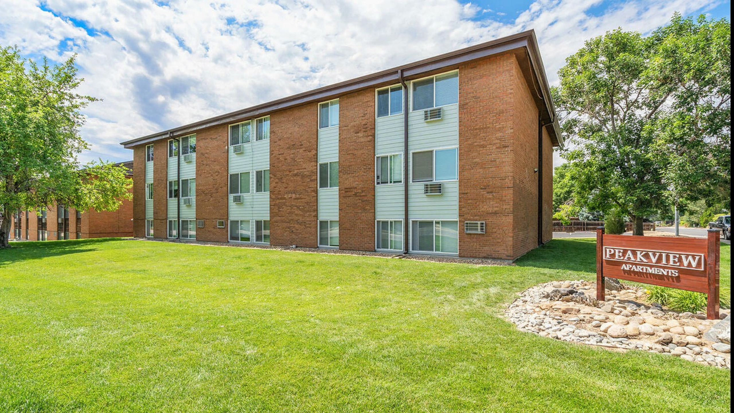 Charming three-story brick apartment building surrounded by lush green lawns and mature trees, with a welcoming Peakview Apartments sign in the foreground under a bright blue sky.