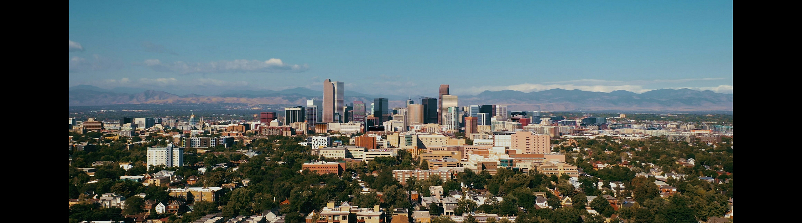Breathtaking panoramic view of a vibrant urban skyline with lush greenery, set against the backdrop of majestic mountains on a clear day.