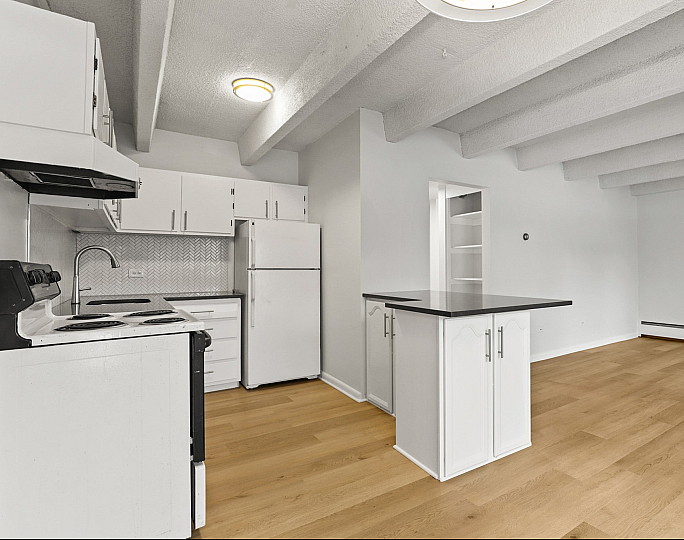 Bright open kitchen with sleek white cabinets and modern black countertops, adjacent to a spacious living area with stylish wooden floors and exposed ceiling beams.