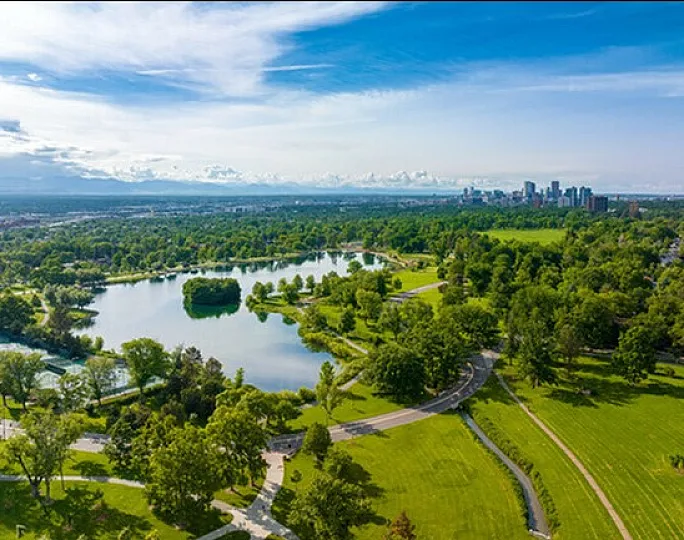Breathtaking aerial view of a lush green park with a serene lake, vibrant trees, and a distant city skyline under a clear blue sky.