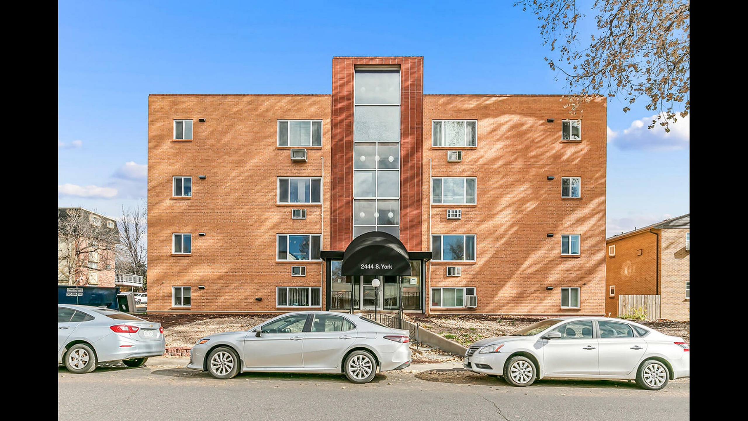 Charming brick apartment building with large windows and a welcoming entrance at 2444 S. York, enhanced by parked cars and a clear blue sky.