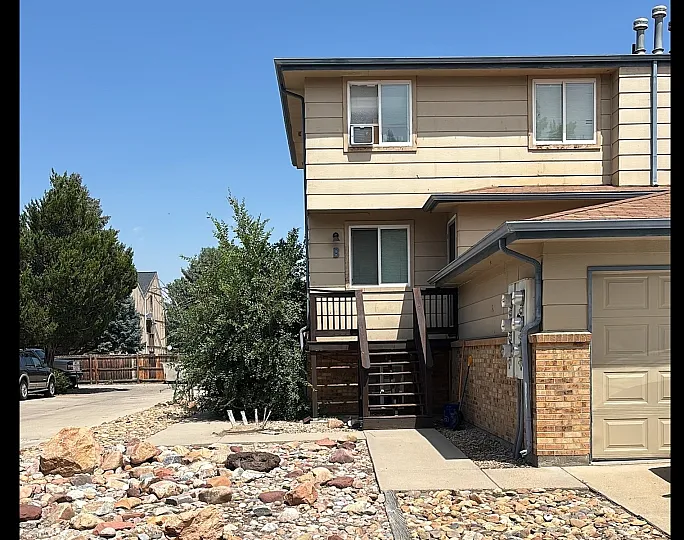 Charming two-story home with rustic landscaping, featuring a rock garden and mature trees under a clear blue sky.