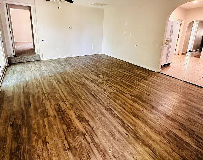 Inviting open-plan living space with elegant wood flooring and modern ceiling fan, leading to a well-lit kitchen area.