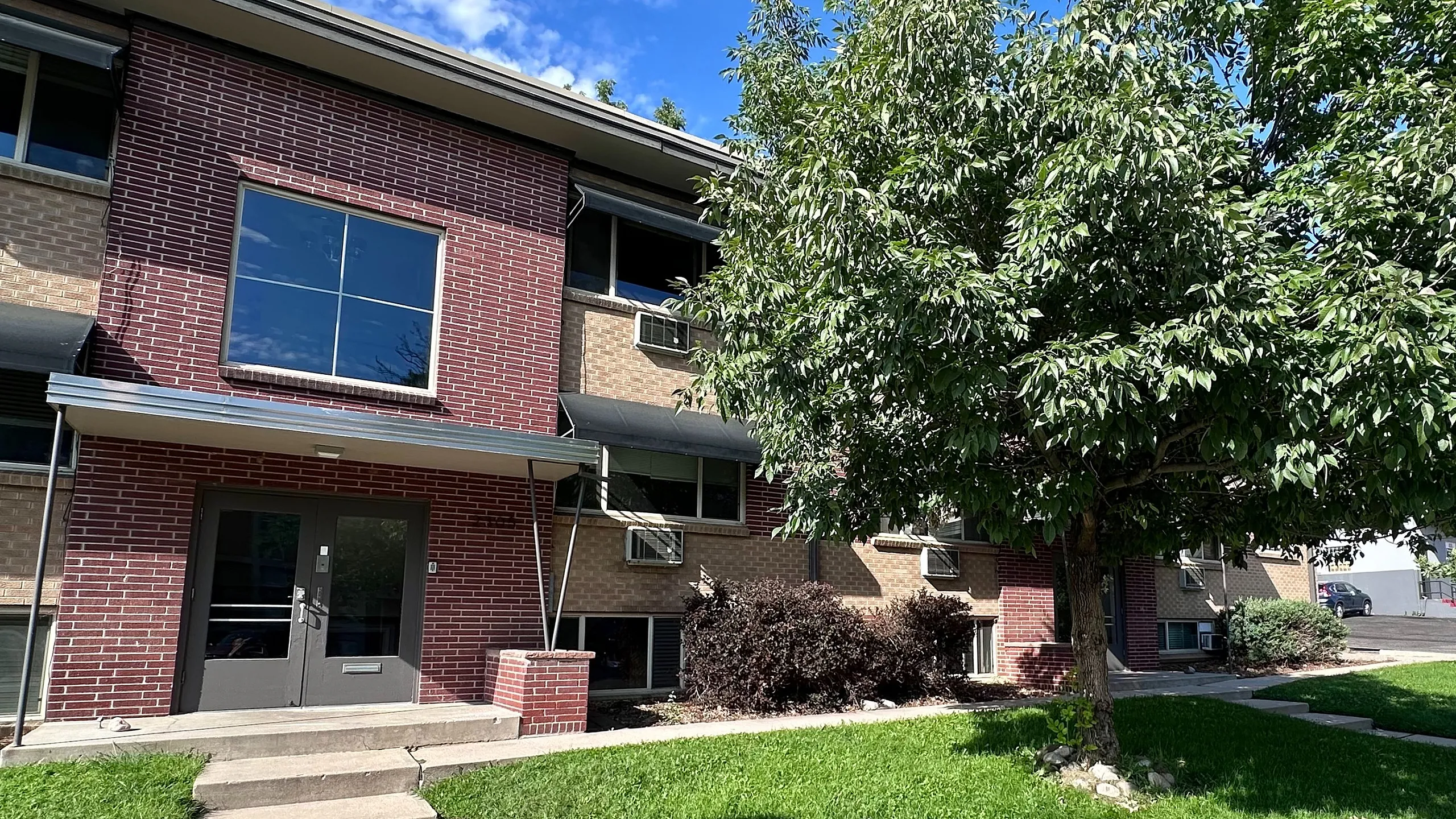 Inviting apartment exterior with charming red brick facade, lush green lawn, and mature tree under a bright blue sky.