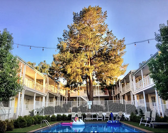 Charming apartment complex with lush greenery, a sparkling pool adorned with fun inflatables, and inviting string lights for a cozy atmosphere.