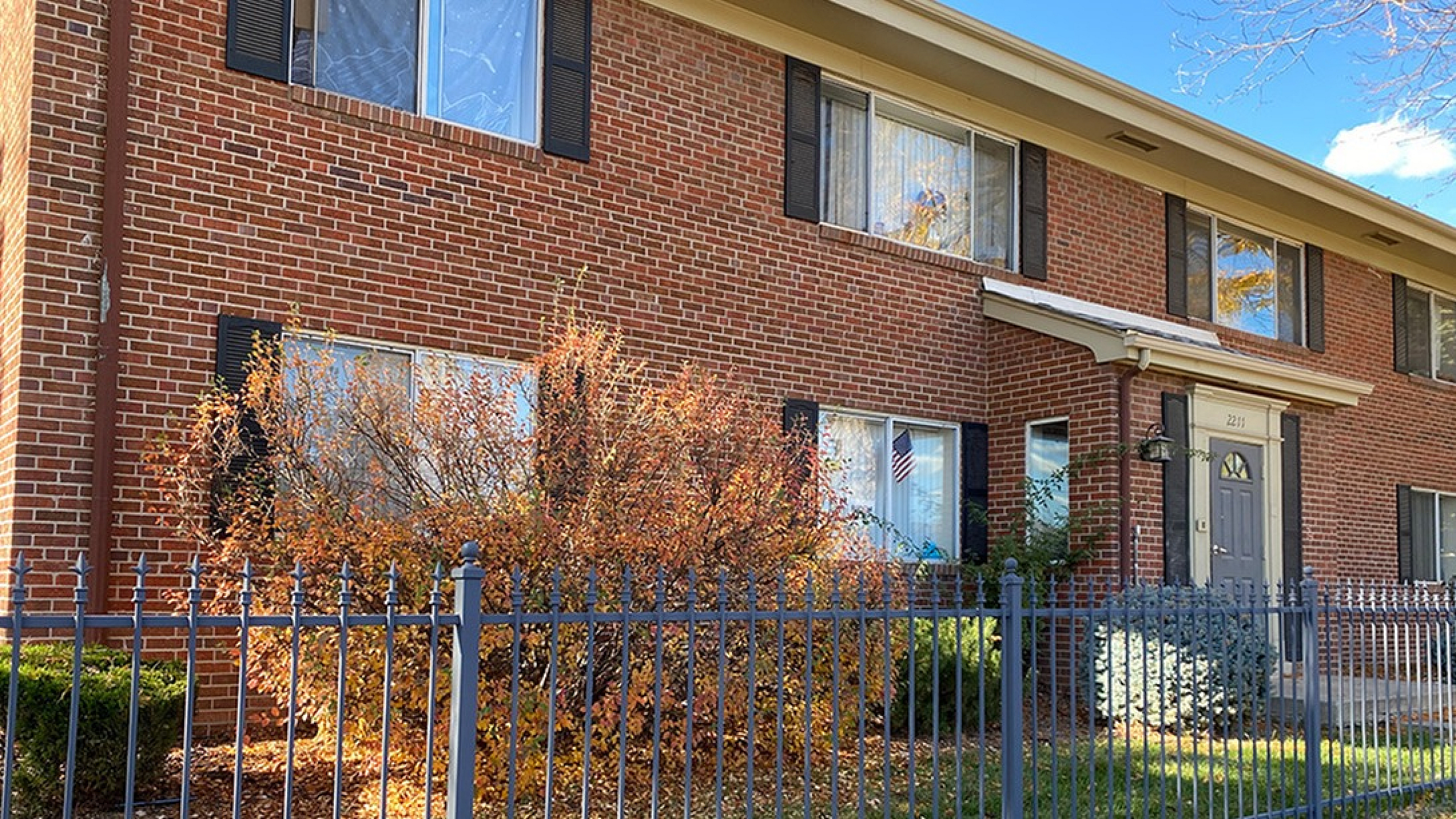 Charming red brick apartment building with large windows, surrounded by a well-manicured garden and a secure metal fence.