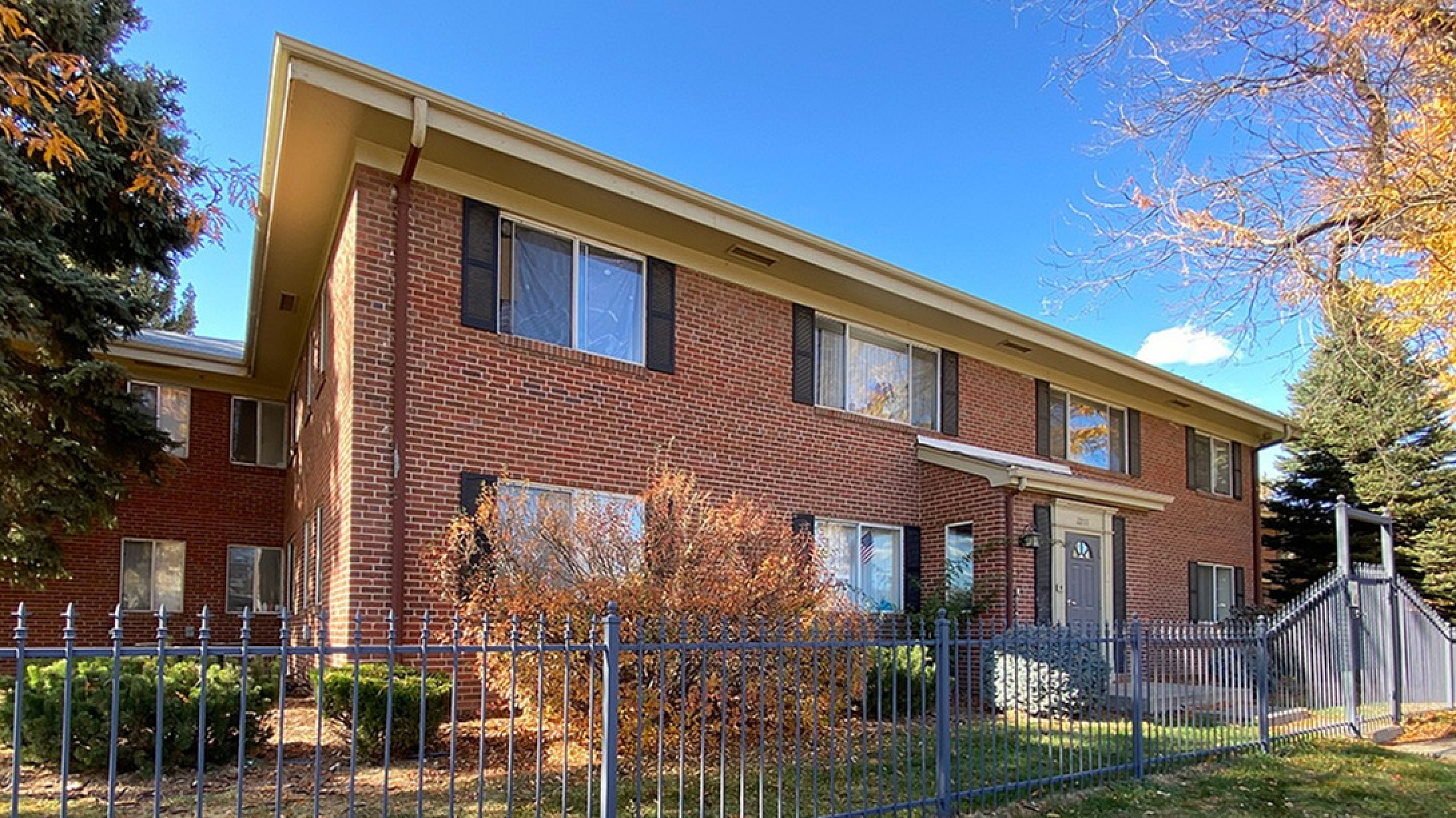 Charming brick apartment building with well-maintained landscaping, surrounded by a decorative fence under a clear blue sky.