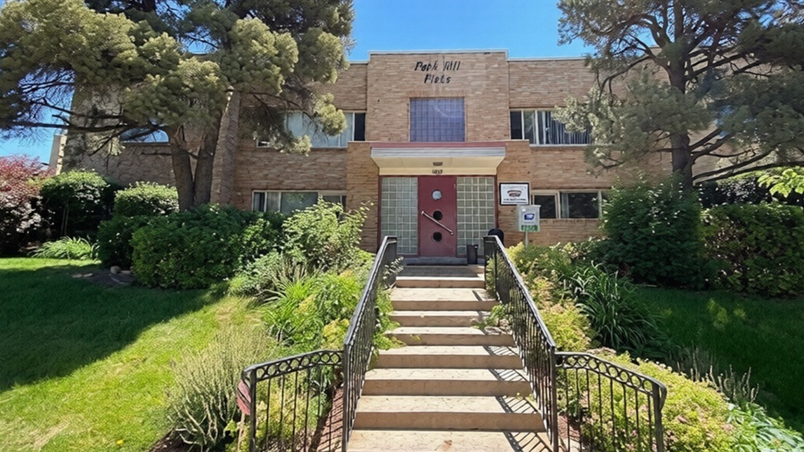 Charming apartment building with a classic brick facade, beautifully surrounded by lush greenery and mature trees, offering a warm and inviting entrance.