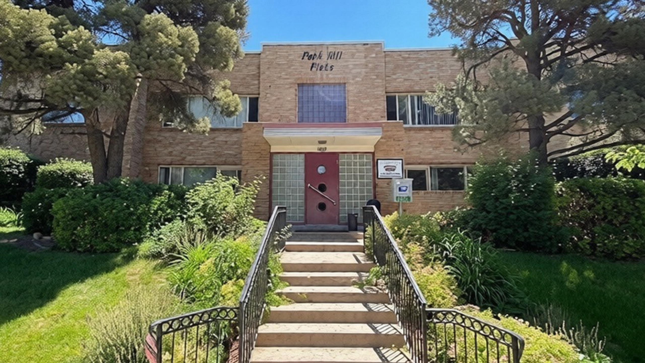 Charming brick apartment building surrounded by lush greenery and vibrant landscaping under a clear blue sky, featuring a welcoming entrance with a classic red door.