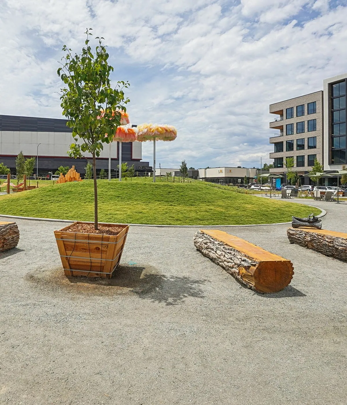 Charming urban park featuring a grassy hill, artistic installations, and modern architecture surrounded by greenery and log-style seating.