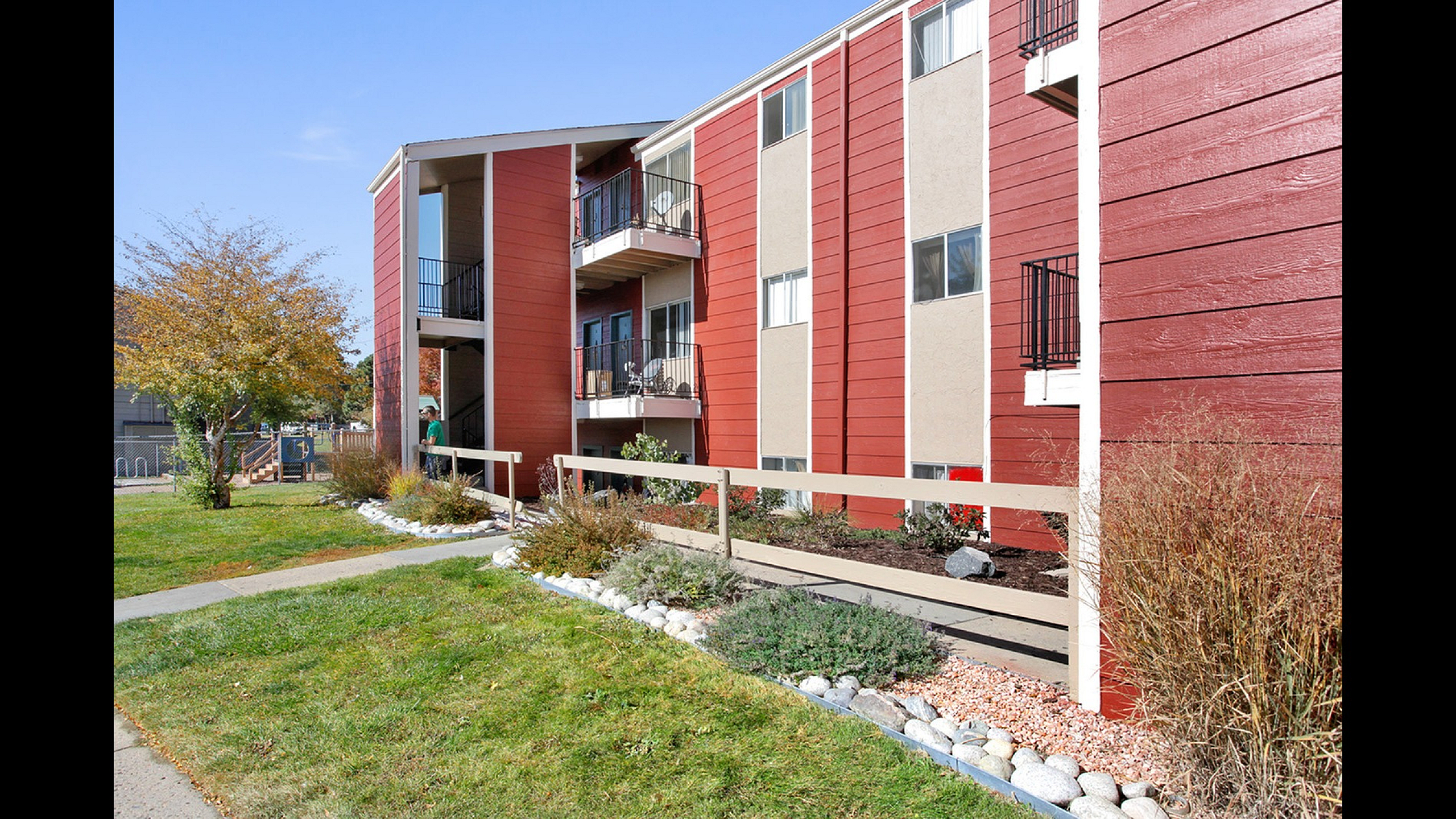 Modern apartment building with vibrant red exterior, featuring balconies and landscaped greenery, offering urban living with a touch of nature.