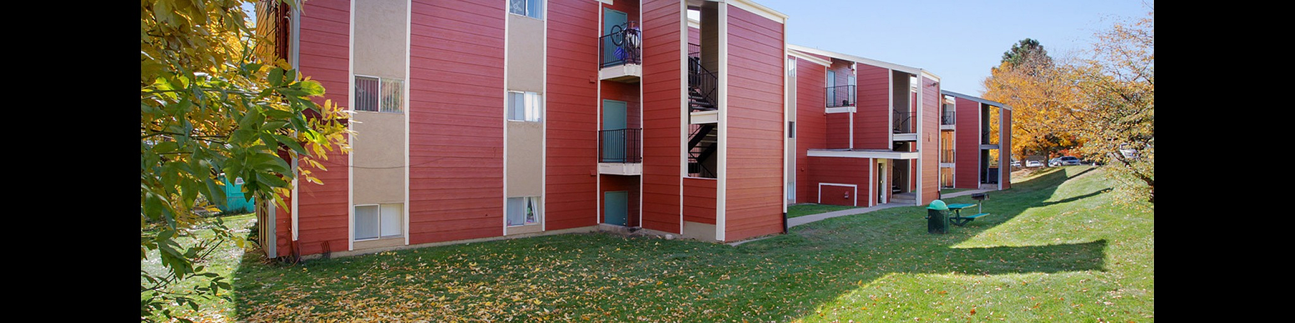 Charming apartment complex exterior with vibrant red siding, surrounded by lush green lawns and autumn foliage, offering serene living.