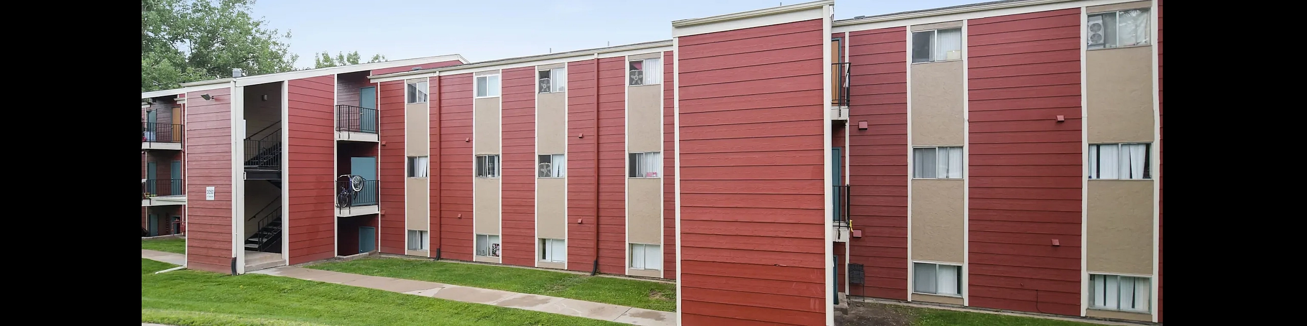 Charming three-story apartment building with vibrant red panels, lush green landscaping, and inviting balconies, perfect for modern living.