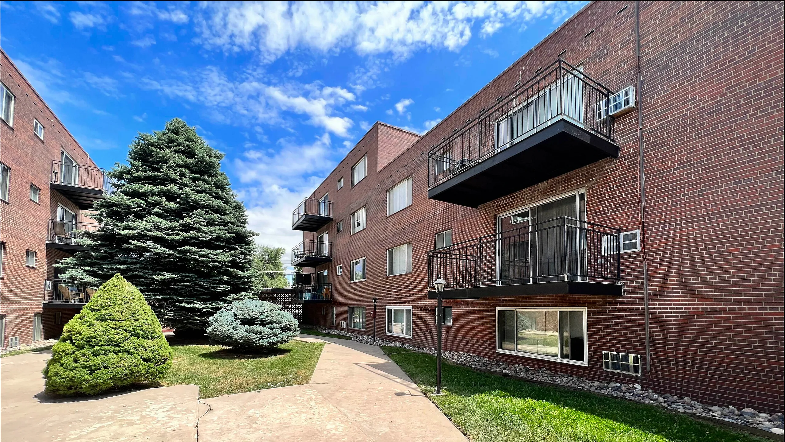Charming brick apartment complex with private balconies, nestled in a serene setting with lush greenery and vibrant blue skies.