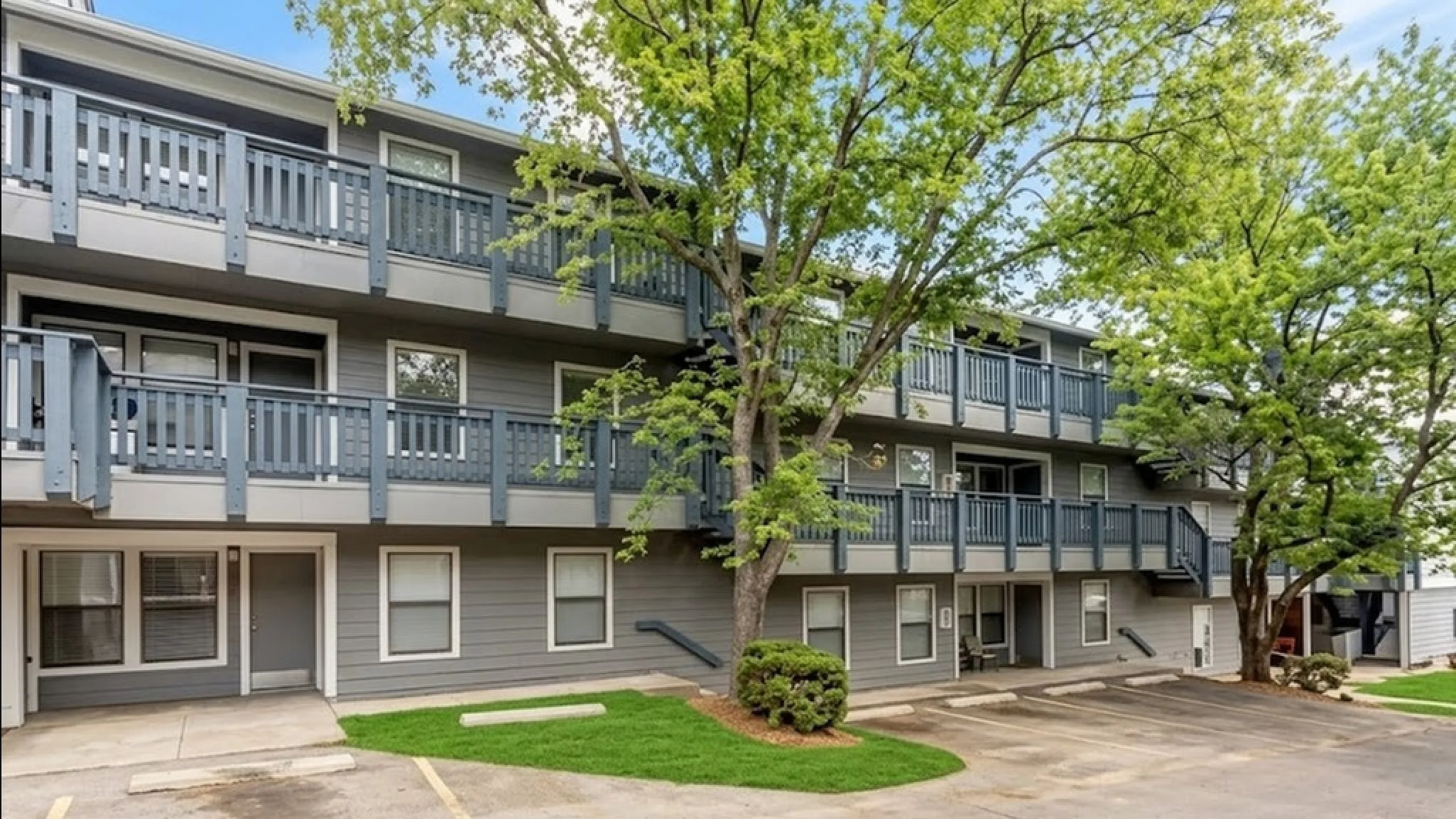 Charming three-story apartment complex with modern gray exterior, surrounded by lush trees and ample parking space.
