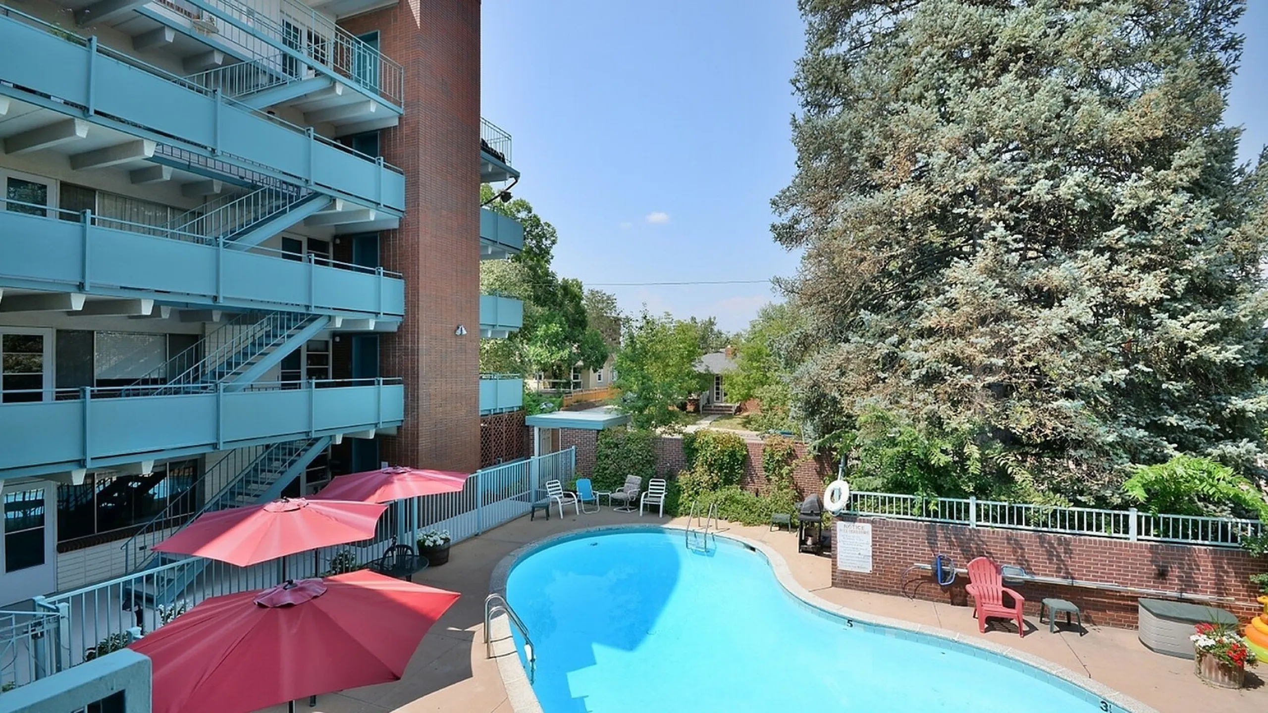 Charming apartment complex with a sparkling outdoor pool, surrounded by lush greenery and vibrant red umbrellas, perfect for relaxation.