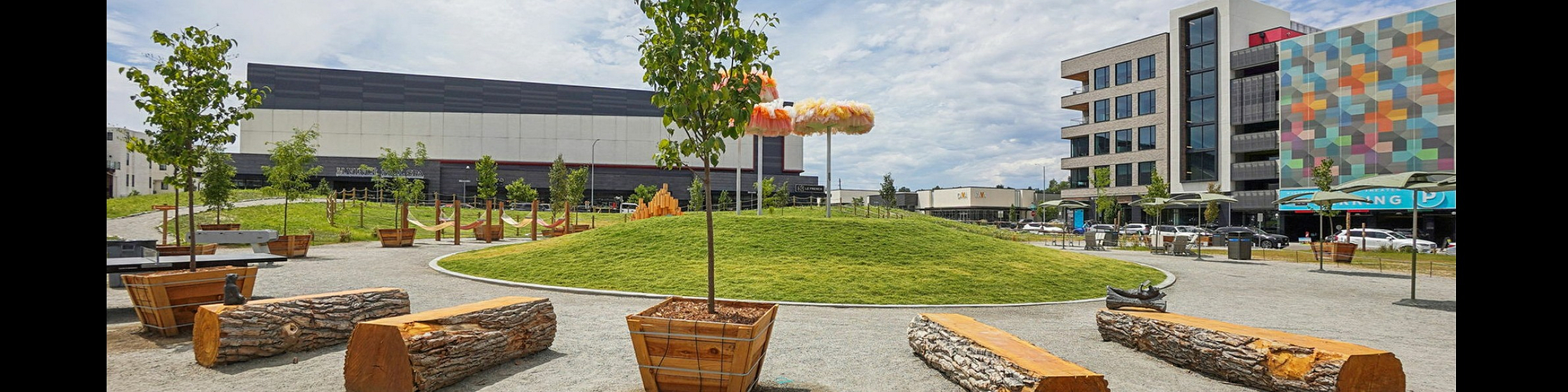 Vibrant urban park setting with lush green grass, modern art installations, and inviting log seating, perfect for relaxation and community gatherings.