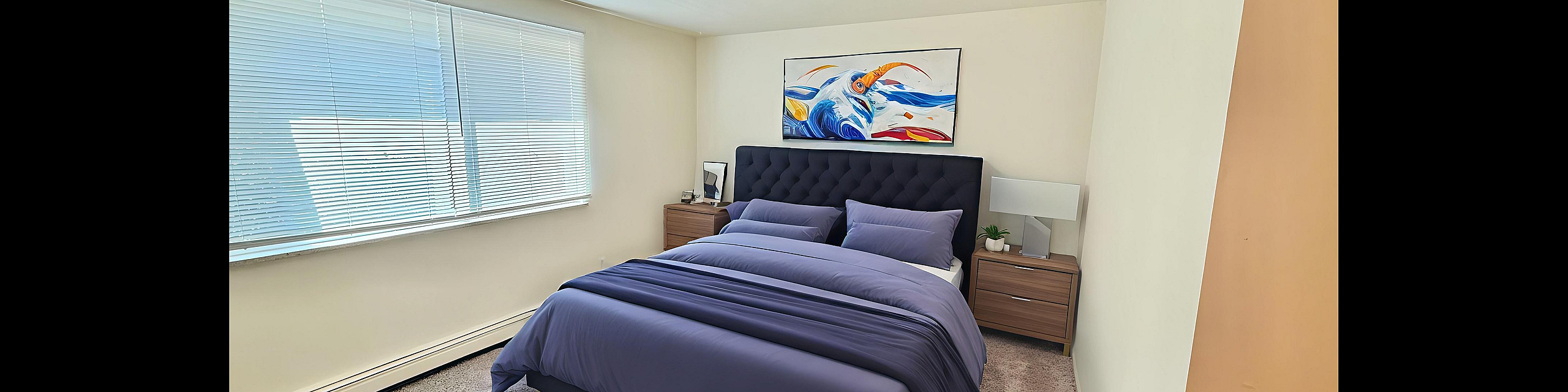 Bright and inviting bedroom with plush carpeting, modern decor, and a cozy bed with vibrant artwork above.