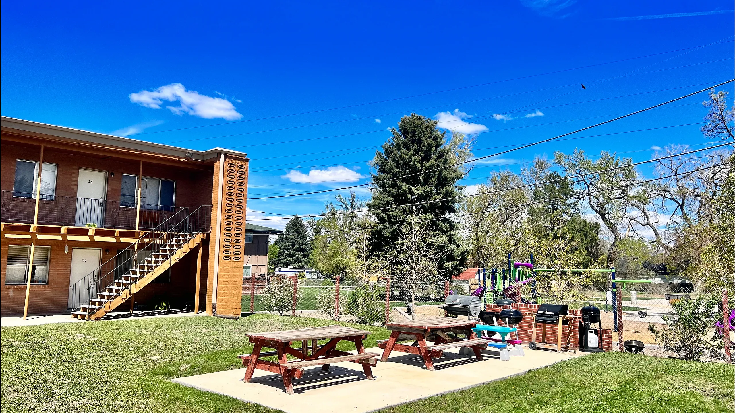 Charming apartment complex with a spacious outdoor area featuring picnic tables, grills, and lush greenery under a clear blue sky.