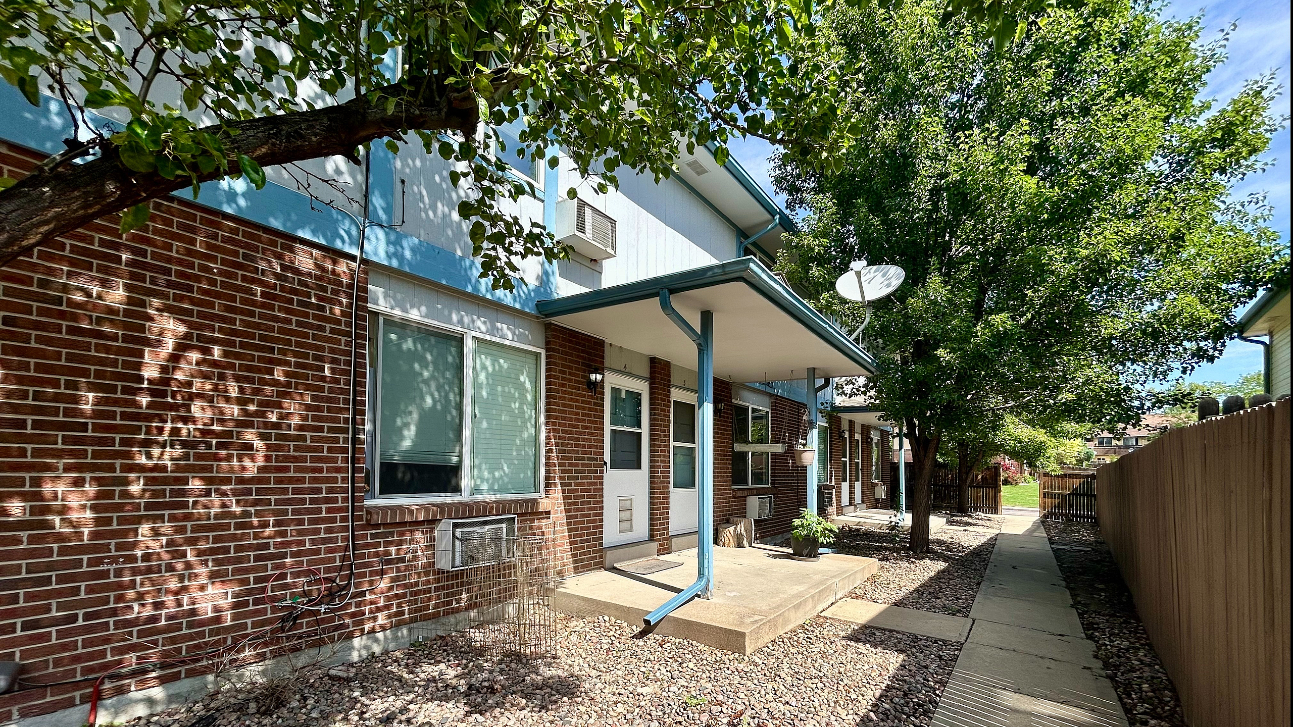 Charming apartment exterior with bright brickwork, shaded by lush trees, and a cozy entryway leading to the serene neighborhood path.