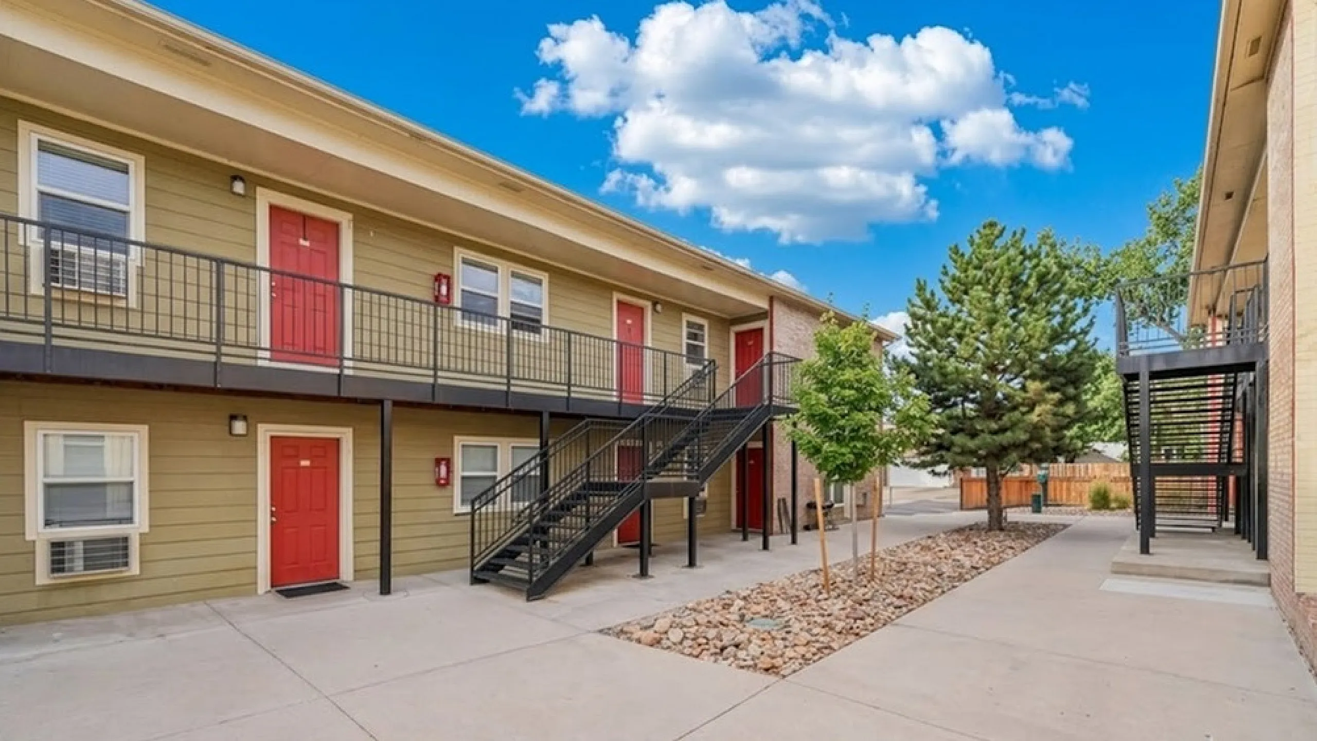 Charming apartment complex featuring vibrant red doors, modern metal staircases, and a well-maintained courtyard with lush greenery and bright blue skies.