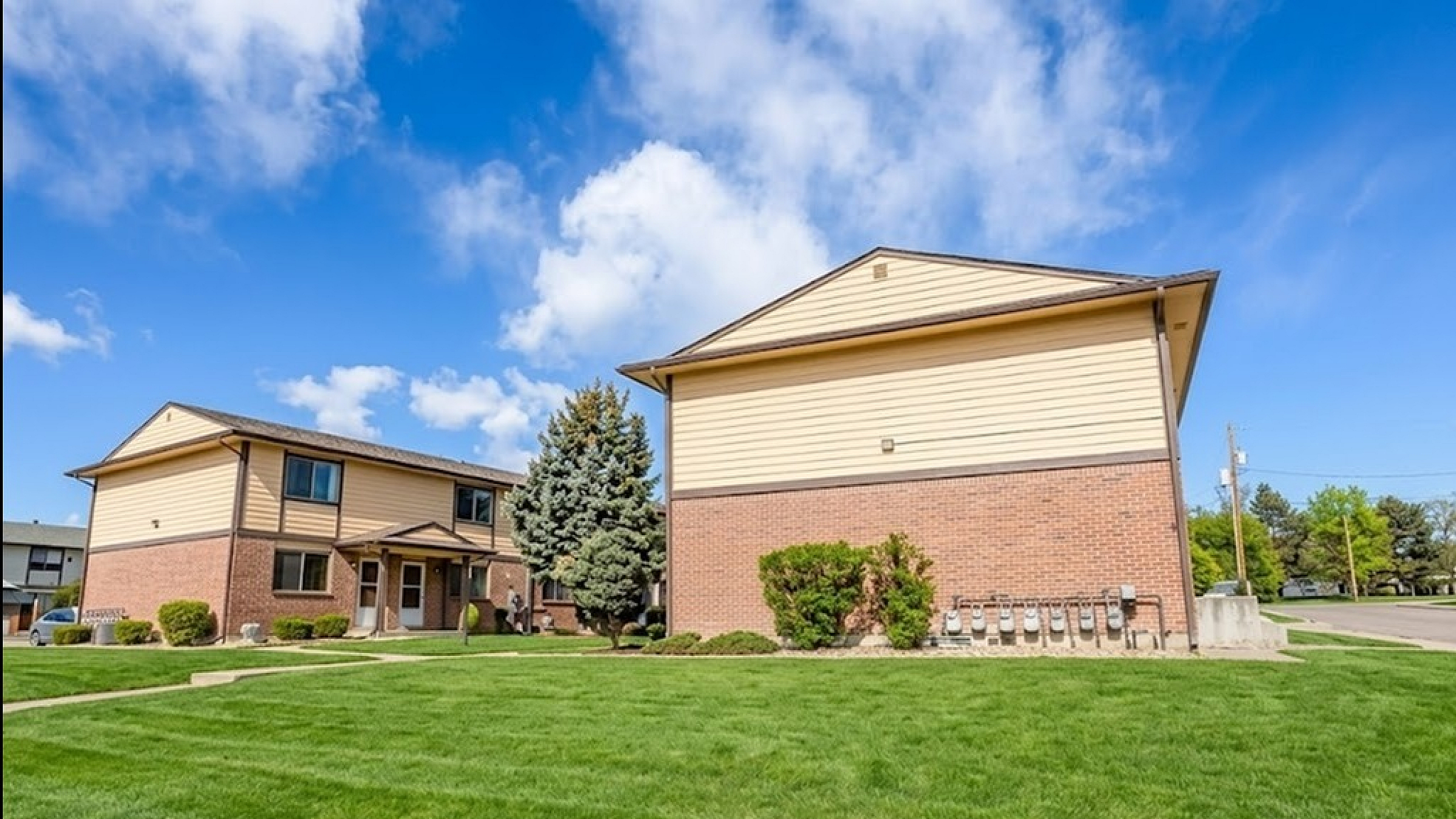 Charming two-story apartment complex with brick and siding exterior, surrounded by manicured green lawns under a bright blue sky.