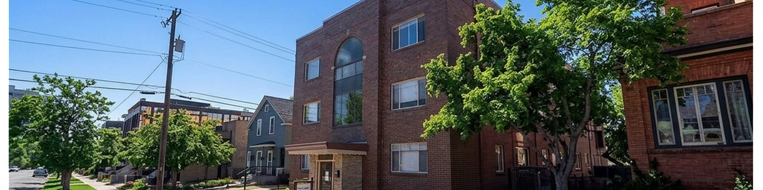 Charming brick apartment building surrounded by lush green lawns and mature trees, set in a picturesque neighborhood on a sunny day.